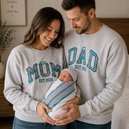 A smiling couple and their newborn baby wearing matching light gray sweatshirts with colorful collegiate-style lettering. The woman's sweatshirt reads 'MOM EST 2026', the man's reads 'DAD EST 2026', and the baby is swaddled in a striped blanket.
