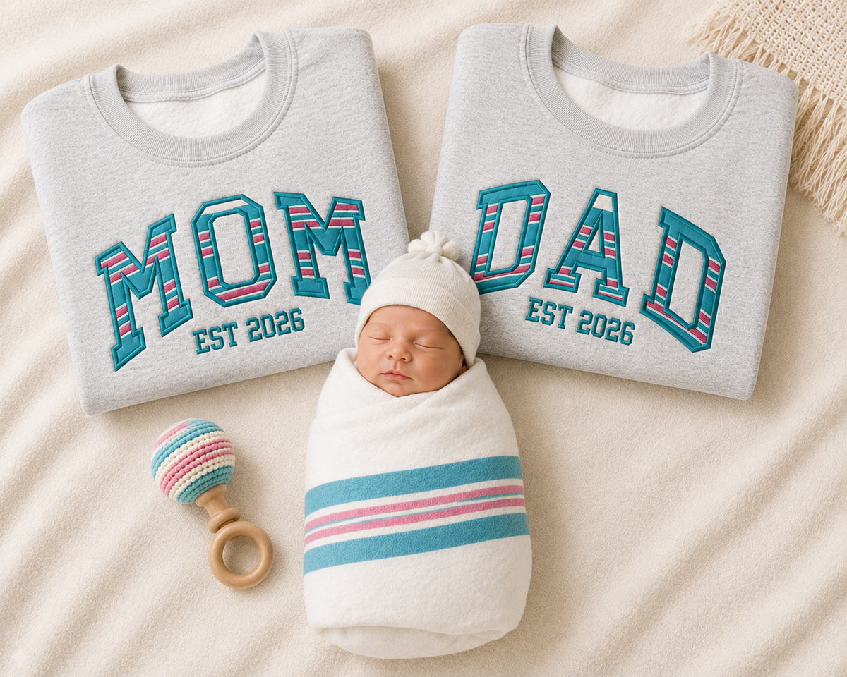 A newborn baby swaddled in white fabric with pink and blue stripes, wearing a white knit hat, posed between two light gray sweatshirts that read 'MOM DAD EST 2026' in teal and pink lettering. A small striped rattle lies to the left on a cream fabric background.